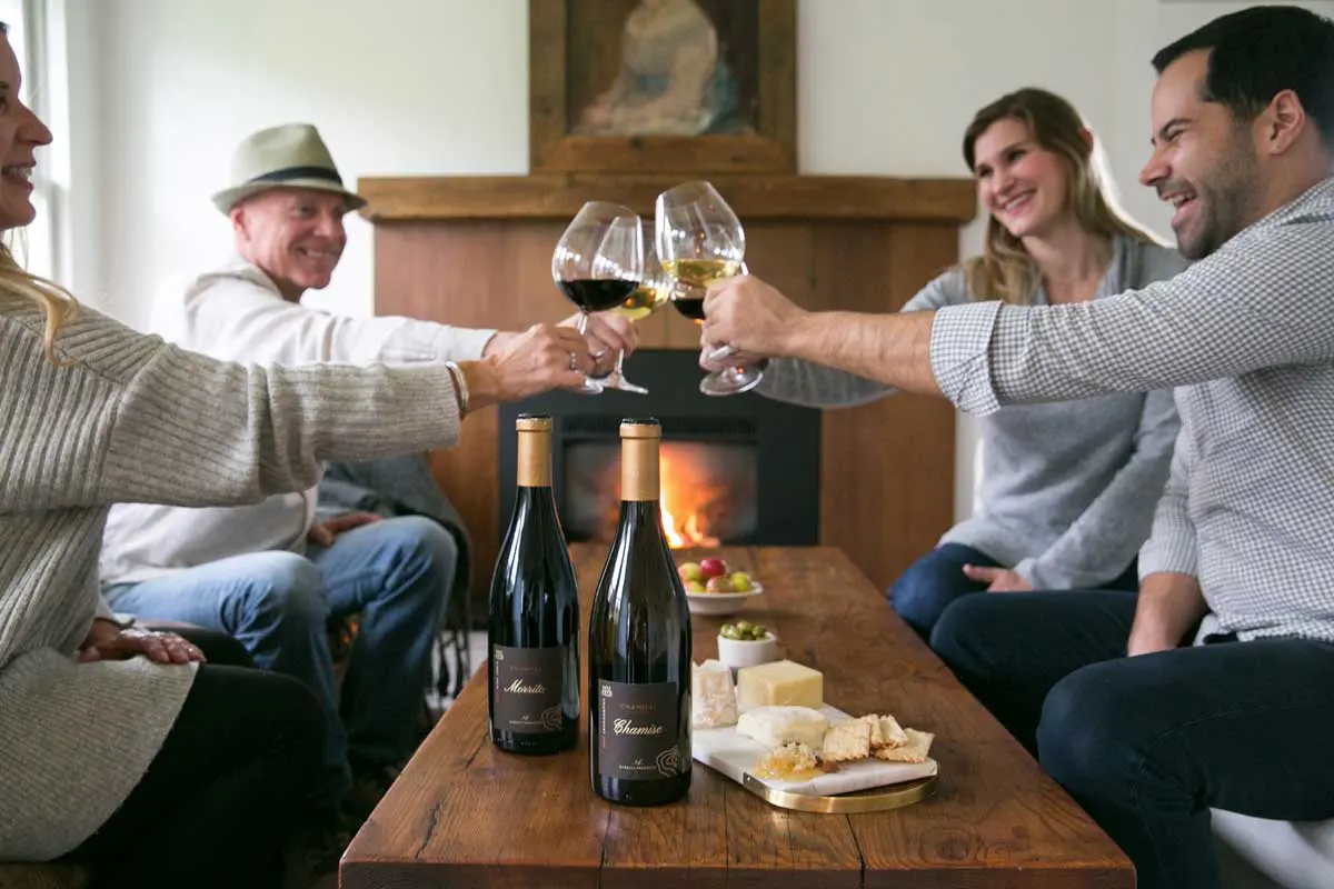 Friends toasting wine glasses beside a fireplace with cheese and wine bottles